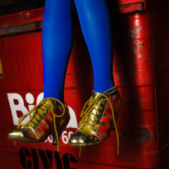 A person wearing electric blue tights and gold laced trainers, sat on a red bin.